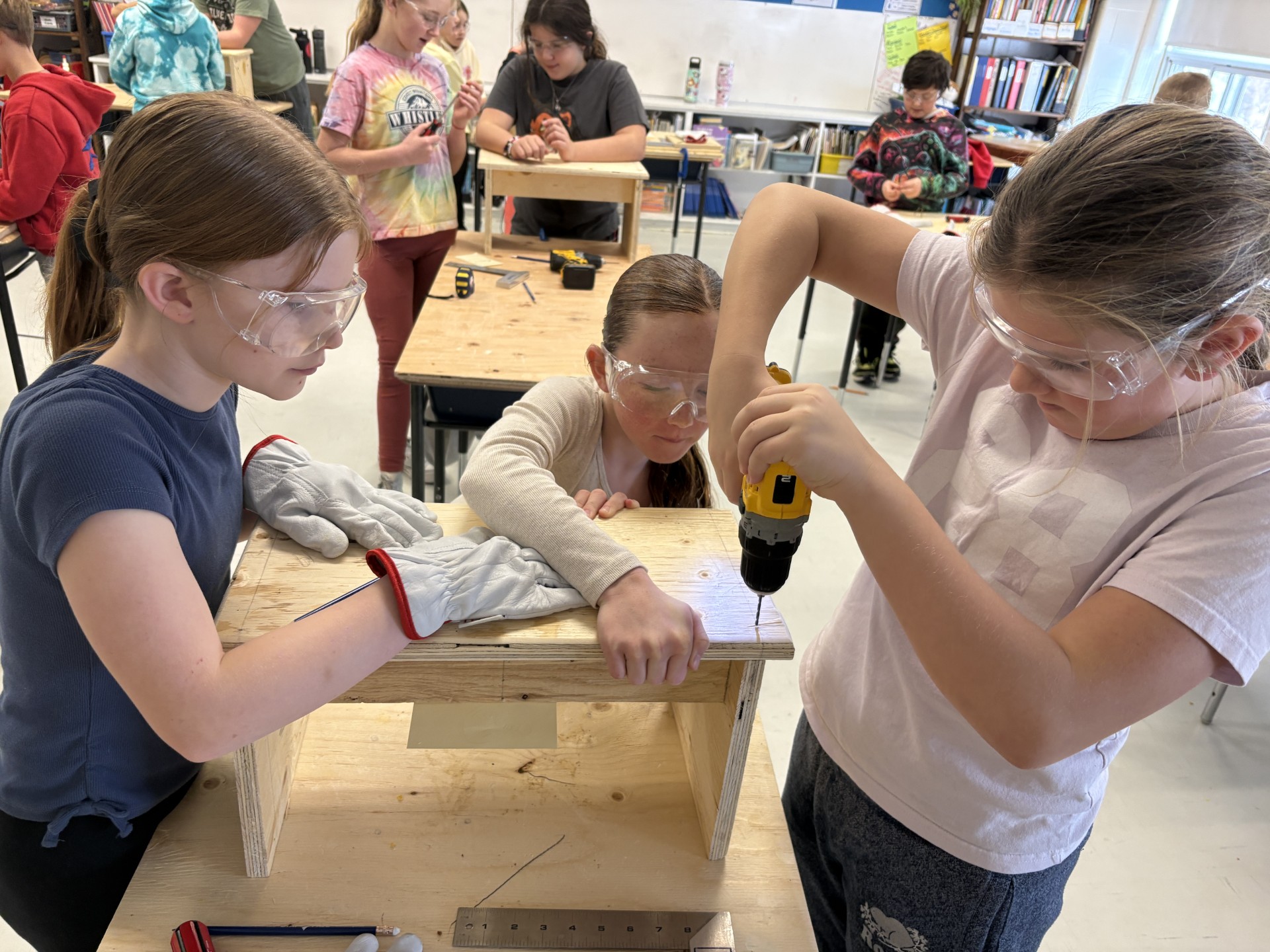 Three students in a classroom working on cookbook stand project. Additional students in the background.