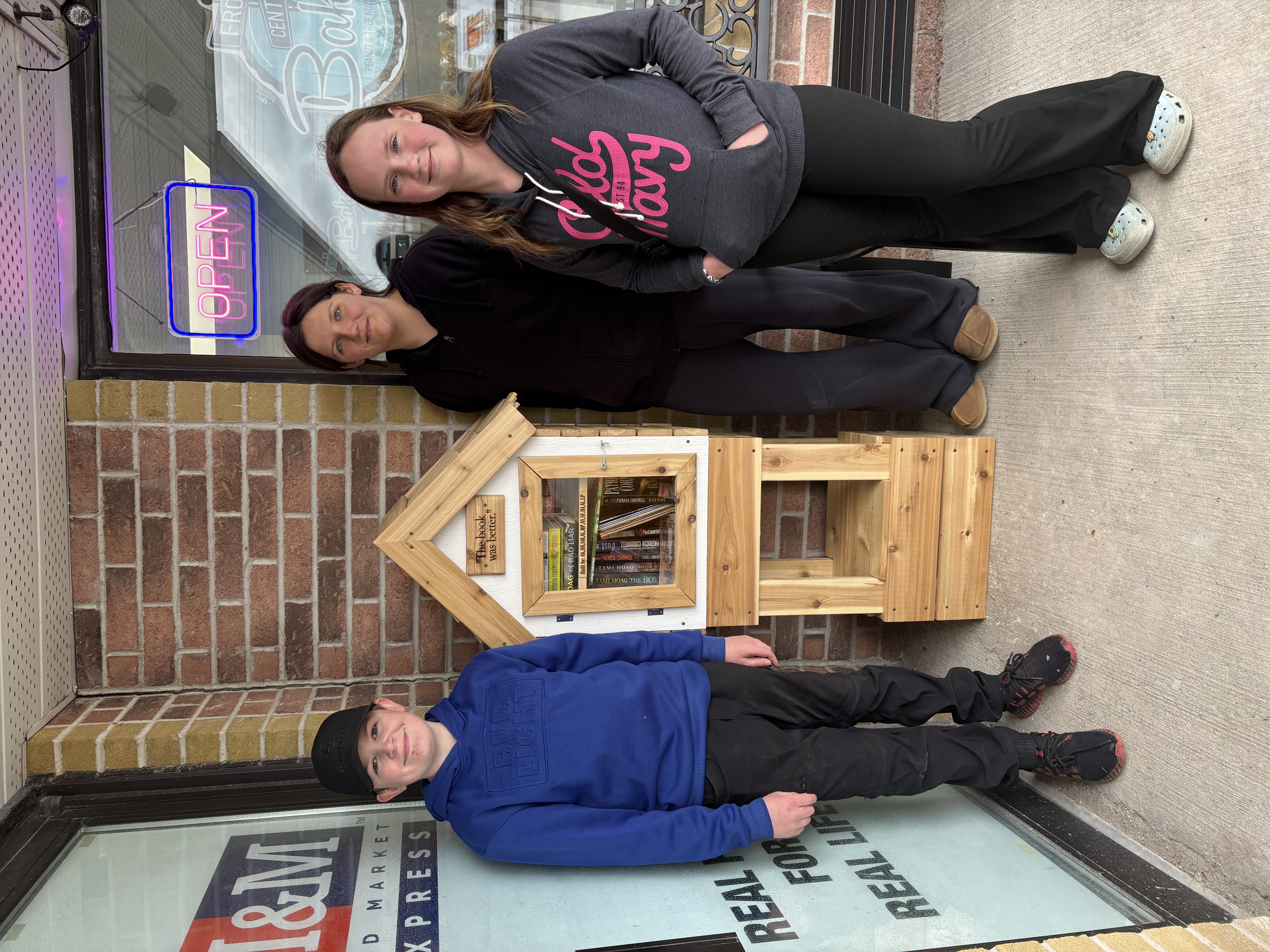 Three Newburgh Public School students stand beside a completed little community library they built, displayed outside of a store.