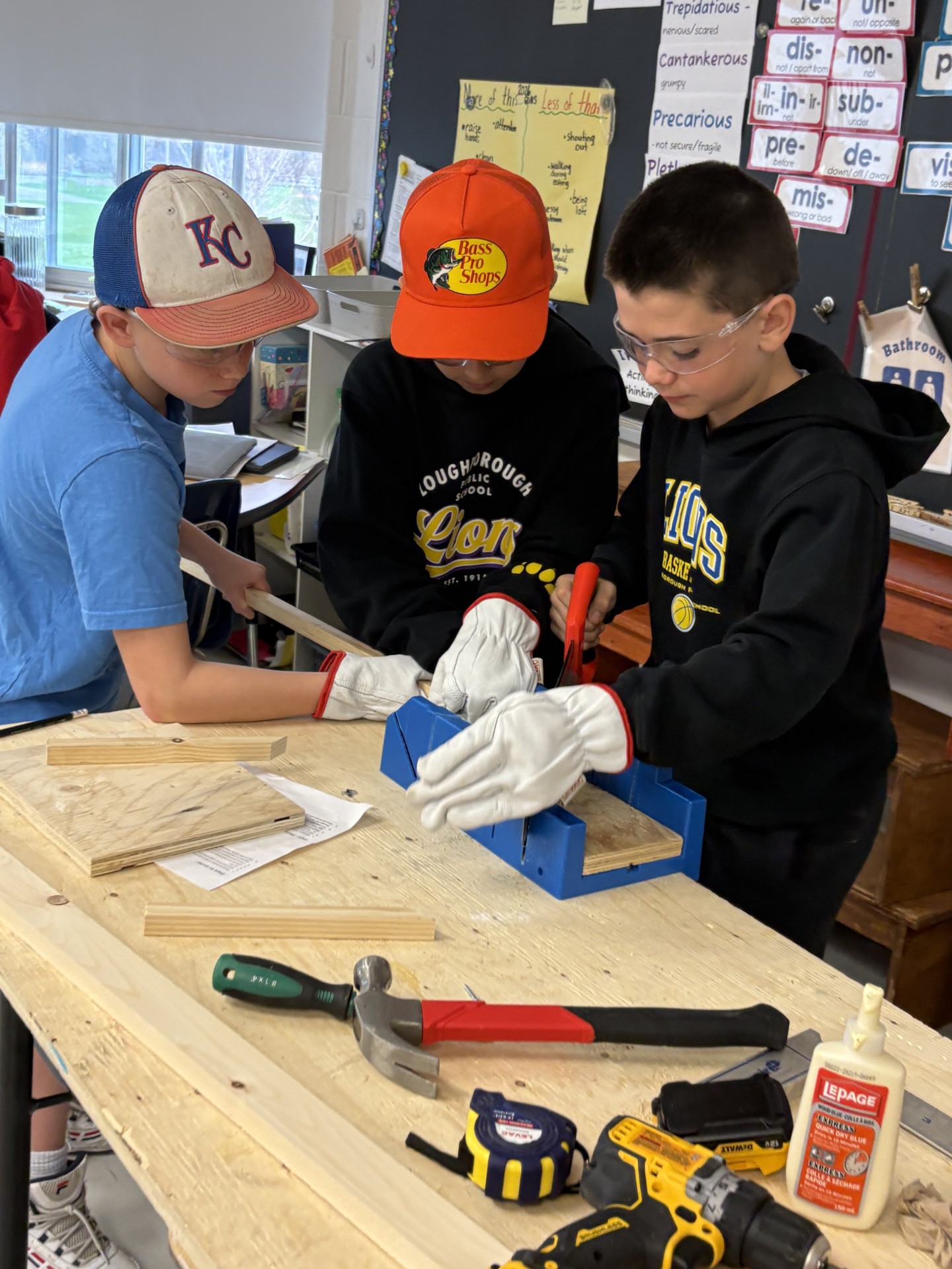 Three students in a classroom working on cookbook stand project. Additional students in the background.