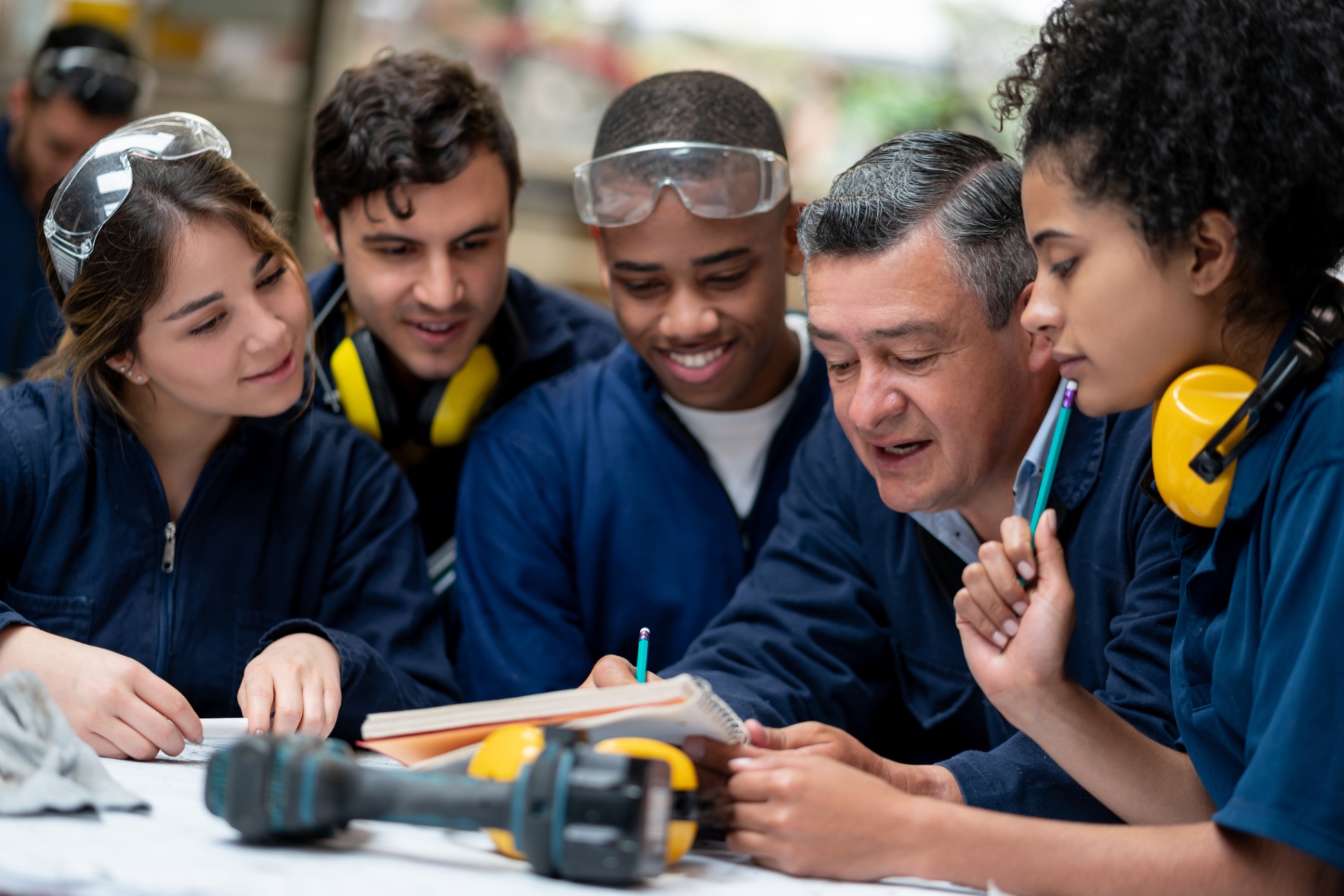 Four students learning with one teacher at a table with safety equipment on and tools on the table