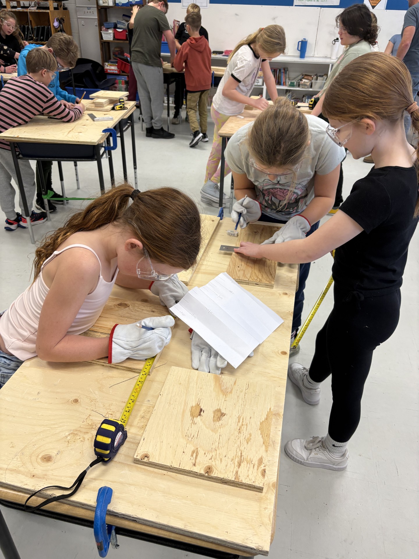 Three students in a classroom working on cookbook stand project. Additional students in the background.