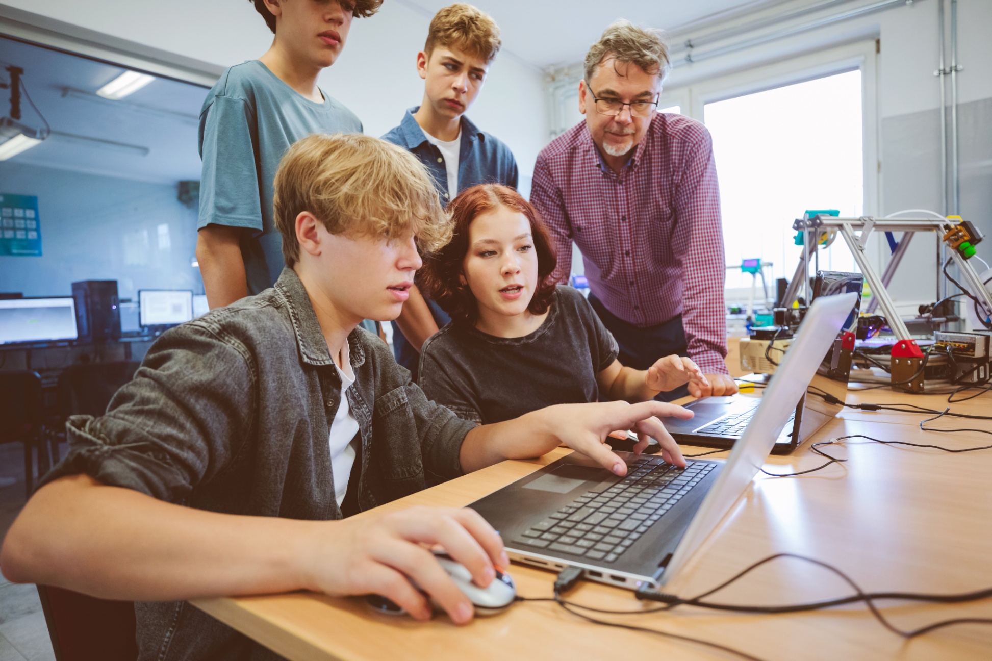Four computer development students learning from a teacher at a table with computers and robotics