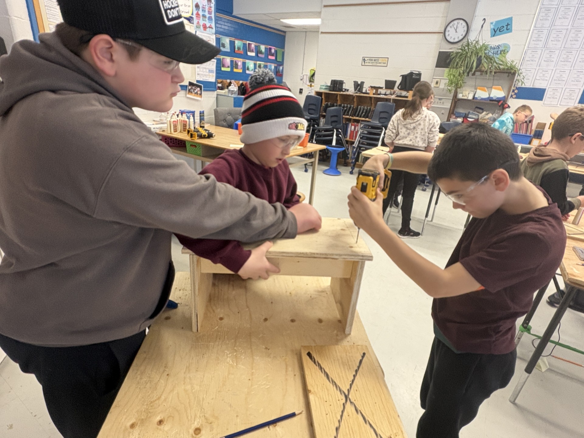 Three students in a classroom working on cookbook stand project. Additional students in the background.