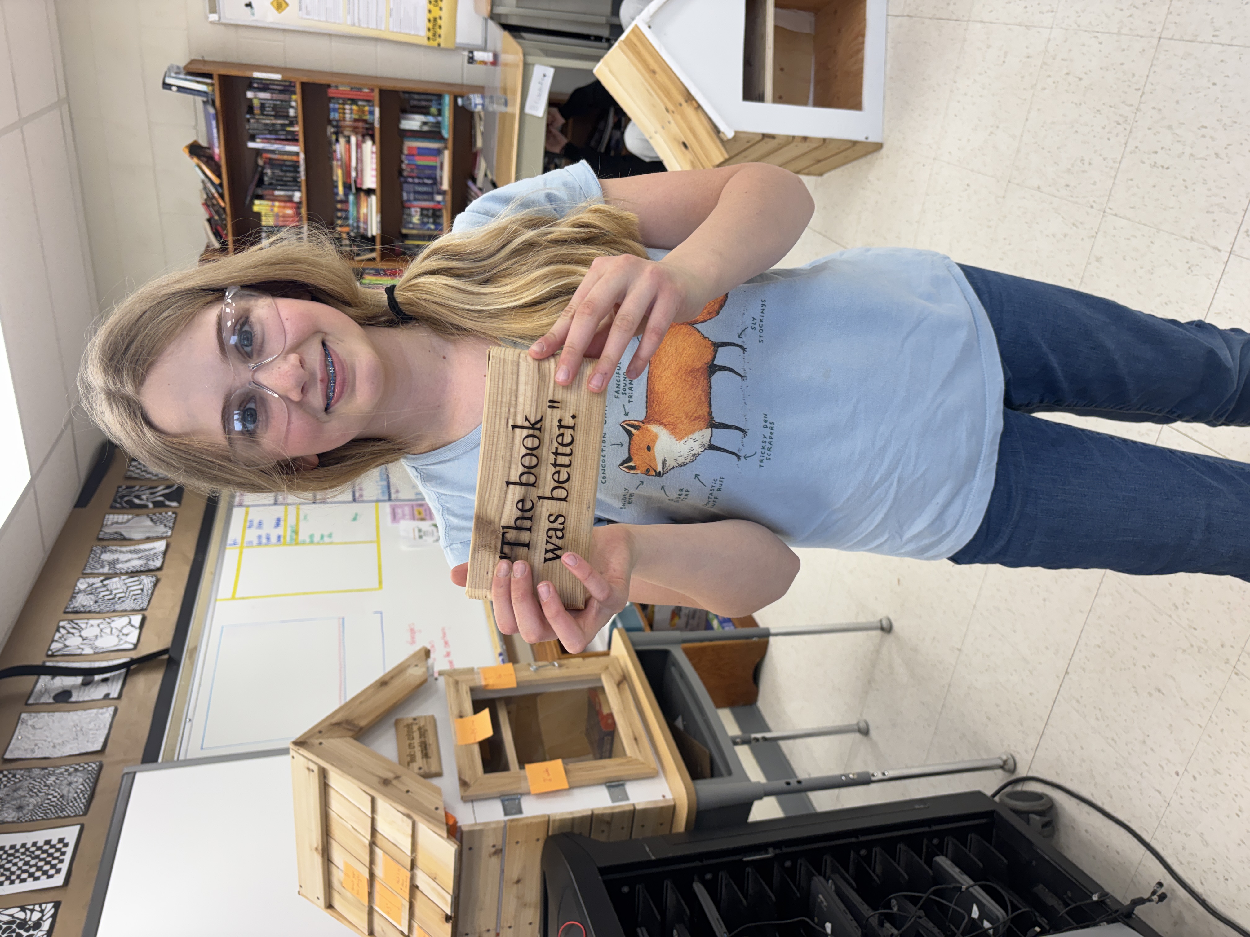 One Newburgh PS student smiles at the camera and holds up a laser-cut plaque qhile working on the little community library project